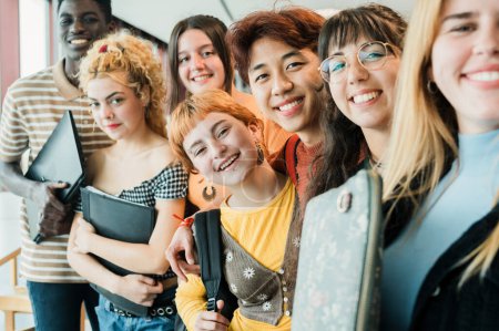 Foto de Grupo de compañeros adolescentes multirraciales felices con computadora portátil y papelería sonriendo y mirando a la cámara mientras toman selfie juntos antes de la lección en la universidad - Imagen libre de derechos