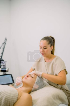 Téléchargez les photos : Esthéticienne utilisant des tissus de visage de cliente contre mur gris au bureau de clinique de cosmétologie - en image libre de droit