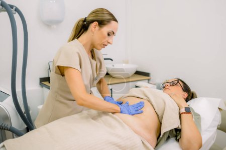 Foto de Esteticista profesional en guantes dando masaje de abdomen para regordete cliente femenino preparándose para adelgazar procedimiento en clínica de belleza - Imagen libre de derechos