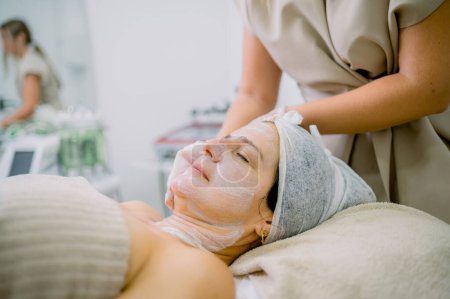 Photo for Crop beautician using tissue to apply rejuvenating cream on cheek of middle aged woman during skin care session in cosmetology clinic - Royalty Free Image