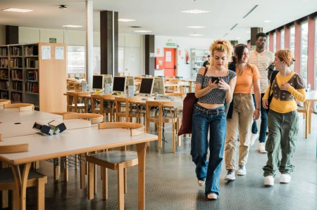 Téléchargez les photos : Groupe de camarades de classe multiraciaux en vêtements décontractés utilisant un smartphone et parlant tout en marchant près des tables dans la bibliothèque lumineuse de l'université moderne - en image libre de droit