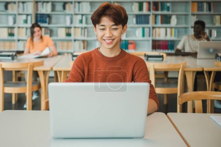 Foto de Feliz adolescente asiático en ropa casual sonriendo y los datos de navegación en netbook mientras está sentado en la mesa y haciendo la tarea de asignación en la biblioteca universitaria - Imagen libre de derechos