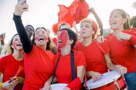 Photo for Delighted young multiethnic female fans in red t shirts with painted faces smiling brightly while taking selfie on smartphone during football match on sunny day - Royalty Free Image