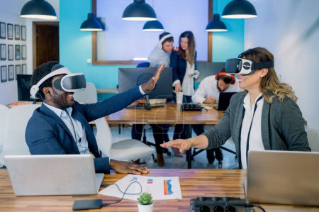 Photo for Positive adult diverse colleagues in formal suits smiling while experiencing virtual reality in VR headset during work in contemporary office - Royalty Free Image