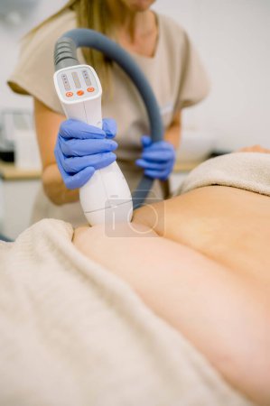 Faceless beautician in gloves applying apparatus of cryolipolysis on abdomen of client with excessive fat
