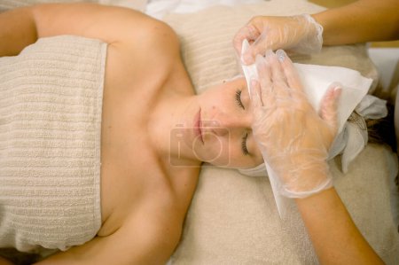Téléchargez les photos : Vue du dessus de l'esthéticienne anonyme dans des gants utilisant une serviette pour essuyer le front de la femme les yeux fermés pendant la séance de soins de la peau à la clinique de cosmétologie - en image libre de droit