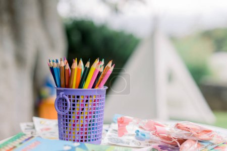 Téléchargez les photos : Lot de crayons multicolores placés à l'intérieur d'une tasse en plastique violet près de la papeterie sur la table dans le jardin - en image libre de droit