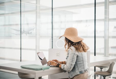 Téléchargez les photos : Vue latérale d'une femme méconnaissable en chapeau de paille assise à table et tapant sur un clavier d'ordinateur portable contre une fenêtre avec un avion volant le jour à l'aéroport - en image libre de droit