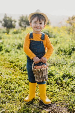 Corps complet d'adorable petit garçon en denim chapeau de paille globale et bottes de gomme jaune souriant et regardant la caméra tout en se tenant debout sur la prairie herbeuse avec panier d'?ufs frais