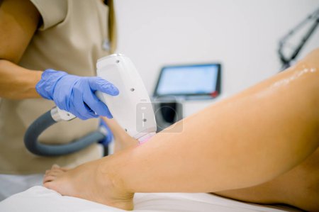 Photo for Faceless beautician in gloves using IPL technology and removing hair from leg of client with cooling gel - Royalty Free Image