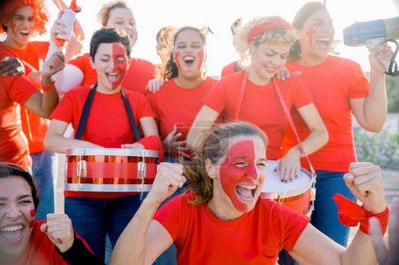 Téléchargez les photos : Contenu jeunes fans féminins multiethniques avec des visages peints en t-shirts rouges serrant les poings et criant joyeusement tout en célébrant la victoire pendant le match de football au coucher du soleil - en image libre de droit