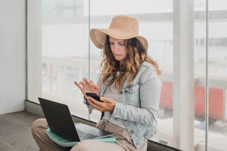 Téléchargez les photos : Jeune femme en vêtements décontractés et chapeau de soleil avec netbook navigation téléphone portable tout en étant assis près de la fenêtre et de travailler à distance dans le terminal de l'aéroport - en image libre de droit