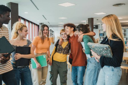 Téléchargez les photos : Asiatique adolescent souriant et embrassant copines près de divers amis tout en se réunissant pour des études dans la bibliothèque universitaire contemporaine - en image libre de droit