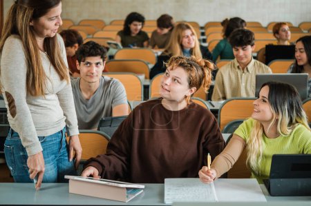 Téléchargez les photos : Enseignante adulte en vêtements décontractés parlant avec des adolescents tout en se tenant près du bureau dans la salle de classe pendant les cours à l'université - en image libre de droit
