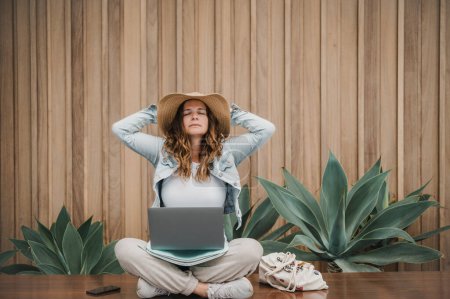Photo for Relaxed mature woman with laptop holding hands behind head and resting with closed eyes while sitting cross legged on border near tropical plants and working on remote project - Royalty Free Image
