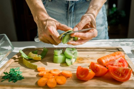 Photo for Crop anonymous woman peeling juicy kiwi with knife on wooden cutting board while preparing ingredients for healthy salad in kitchen - Royalty Free Image