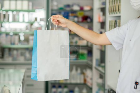 Photo for Side view of crop anonymous female pharmacist in white uniform and protective mask giving paper bag with personalized pills to customer at drugstore - Royalty Free Image