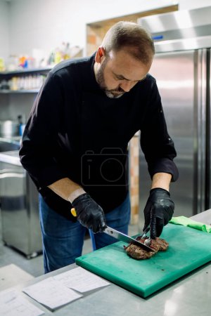 Photo for Bearded focused male chef in casual shirt and jeans cutting piece of delicious roasted meat over cutting board while working in modern restaurant - Royalty Free Image