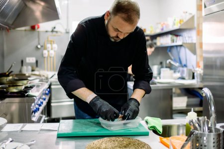 Foto de Chef profesional hombre barbudo en guantes negros acusar a los alimentos en un recipiente transparente mientras trabaja en la cocina profesional - Imagen libre de derechos
