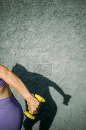 Foto de Vista lateral de la hembra deportiva sin rostro en ropa deportiva sosteniendo la mancuerna amarilla y haciendo kickback tríceps contra la pared gris - Imagen libre de derechos