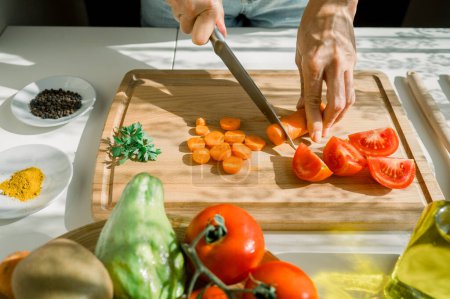 Foto de Alto ángulo de cultivo mujer anónima corte zanahoria en tabla de cortar de madera cerca de tomates y perejil en la cocina durante el fin de semana soleado - Imagen libre de derechos