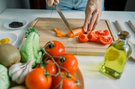 Foto de Alto ángulo de cultivo mujer anónima rebanando zanahoria fresca en la tabla de cortar mientras se prepara ensalada con tomates y perejil en la cocina - Imagen libre de derechos