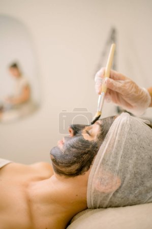 Photo for Relaxed female client with closed eyes lying on table while crop cosmetologist applying carbon mask with brush in beauty salon - Royalty Free Image