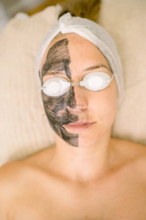 Foto de Vista superior del cliente femenino en gafas protectoras con máscara de carbono en la mitad de la cara durante la sesión de cuidado de la piel en el salón - Imagen libre de derechos