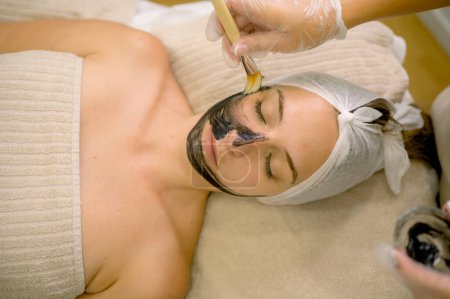 Téléchargez les photos : Vue du dessus de l'esthéticienne des cultures à l'aide d'une brosse pour répandre le masque de carbone sur le visage de la femme aux yeux fermés pendant la procédure de soins de la peau dans le salon de cosmétologie - en image libre de droit