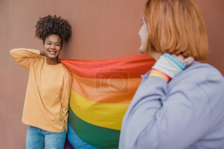Photo for Happy multiracial girlfriends smiling and looking at each other while carrying rainbow flag against brown wall - Royalty Free Image