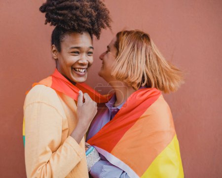 Foto de Feliz pareja de lesbianas multiétnicas sonriendo y abrazándose bajo la bandera LGBT contra el fondo marrón - Imagen libre de derechos