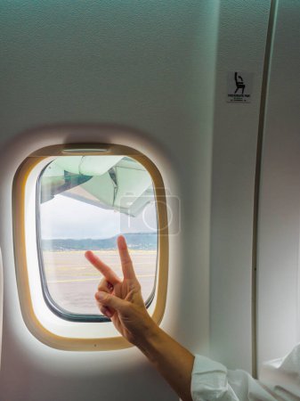 Foto de Mano de cultivo turista irreconocible que muestra gesto de paz mientras está sentado en el asiento del pasajero cerca del iluminador durante el vuelo en aviones modernos - Imagen libre de derechos