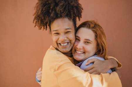 Téléchargez les photos : Vue latérale de filles adolescentes multiraciales heureuses s'embrassant et regardant la caméra avec le sourire sur fond brun - en image libre de droit
