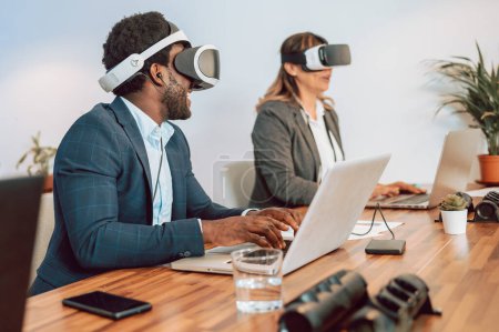 Foto de Colegas en gafas VR modernas navegando netbooks en la mesa mientras exploran juntos la realidad virtual durante la jornada laboral en la oficina moderna de luz - Imagen libre de derechos