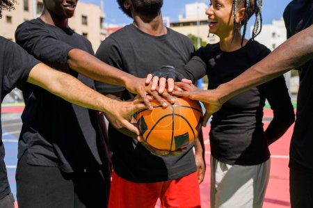 Téléchargez les photos : Cultivez les joueurs anonymes afro-américains de basket-ball en vêtements de sport toucher la balle et mettre les mains ensemble tout en se tenant debout sur un terrain de sport coloré avant l'entraînement le jour de l'été - en image libre de droit