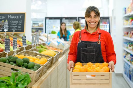Téléchargez les photos : Femme hispanique positive en chemise rouge et tablier souriant et regardant la caméra tout en portant une caisse en bois avec des oranges fraîches pendant le travail dans un supermarché - en image libre de droit