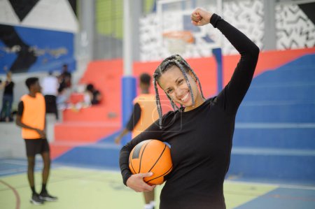Téléchargez les photos : Joueuse afro-américaine excitée de basket-ball debout avec la balle sur le terrain de jeu et célébrer la victoire dans le jeu avec le poing serré tout en regardant la caméra - en image libre de droit