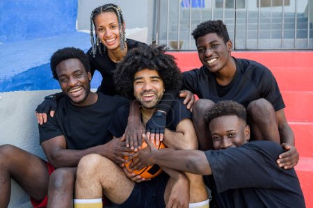 Foto de Grupo de personas afroamericanas encantadas en ropa deportiva tocando el baloncesto y mirando a la cámara mientras están sentados en las escaleras durante el entrenamiento en la calle - Imagen libre de derechos