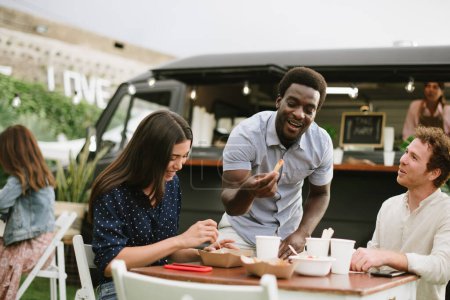 Foto de Feliz hombre negro tomando chip con ketchup y sonriendo mientras almuerza con amigos cerca de camión de comida en el día - Imagen libre de derechos
