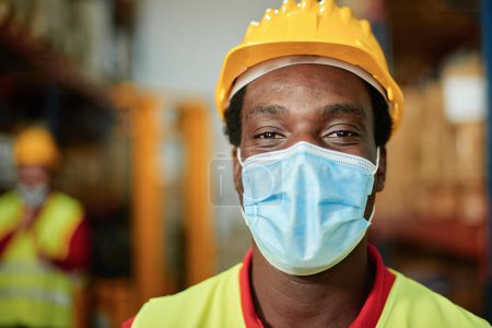 Téléchargez les photos : Portrait d'un ouvrier africain à l'intérieur d'un entrepôt avec masque de sécurité - Focus sur les yeux des hommes - en image libre de droit