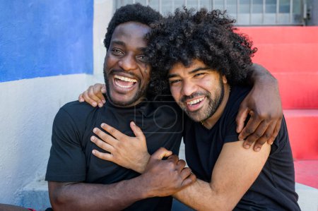 Téléchargez les photos : Amis masculins afro-américains positifs en vêtements de sport embrassant tout en étant assis sur des marches colorées pendant l'entraînement sur la rue de la ville - en image libre de droit