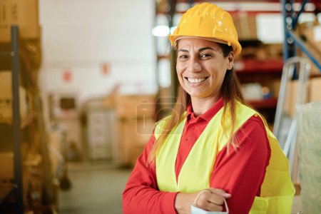 Foto de Retrato de una mujer trabajadora caucásica adulta feliz mirando a la cámara dentro de un almacén con un sombrero duro y ropa de seguridad - Enfoque en la cara - Imagen libre de derechos
