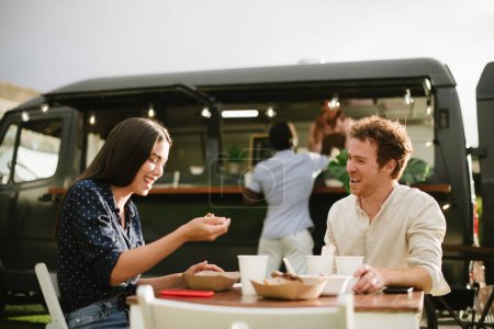 Téléchargez les photos : Un homme et une femme joyeux en vêtements décontractés riant de plaisanterie assis à table et mangeant des plats à emporter près du van - en image libre de droit
