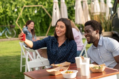 Photo for Cheerful multiracial male and female friends taking self shot while sitting at table with takeaway food in outdoors cafe - Royalty Free Image