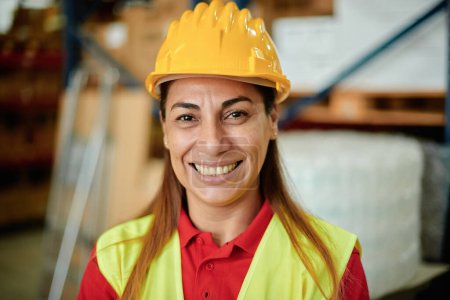 Foto de Retrato de una mujer trabajadora caucásica adulta feliz mirando a la cámara dentro de un almacén con un sombrero duro y ropa de seguridad - Enfoque en la cara - Imagen libre de derechos