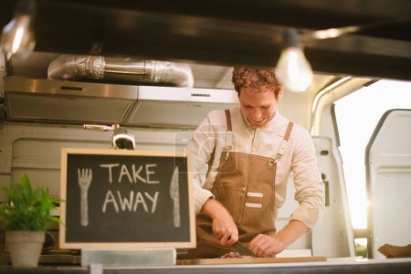 Foto de Bajo ángulo de alegre chef masculino en camisa blanca y delantal sonriendo y picando ingredientes cerca de pizarra con inscripción para llevar durante el trabajo en camión de comida - Imagen libre de derechos
