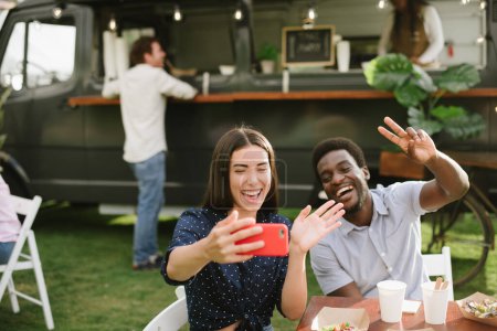Foto de Mujer alegre saludando de la mano y tomando selfie con novio negro gesto V signo mientras almuerza en el patio cerca de camión de comida - Imagen libre de derechos