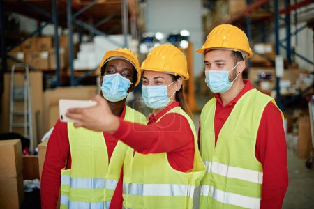 Photo for Male engineer in protective uniform and protective mask taking self portrait on smartphone while working in warehouse - Royalty Free Image