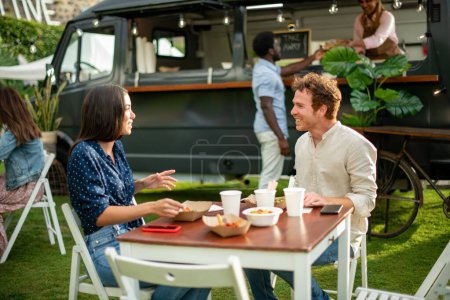 Foto de Pareja positiva sentada en la mesa con comida para llevar y hablando entre sí contra camión de comida en el parque de verano - Imagen libre de derechos
