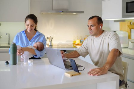 Photo for Positive mother holding infant while sitting at table near husband making video call via laptop in modern kitchen - Royalty Free Image